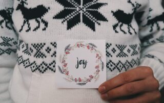 Close up of a man in a white Christmas jumper holding a Christmas card that has the word Joy on it.