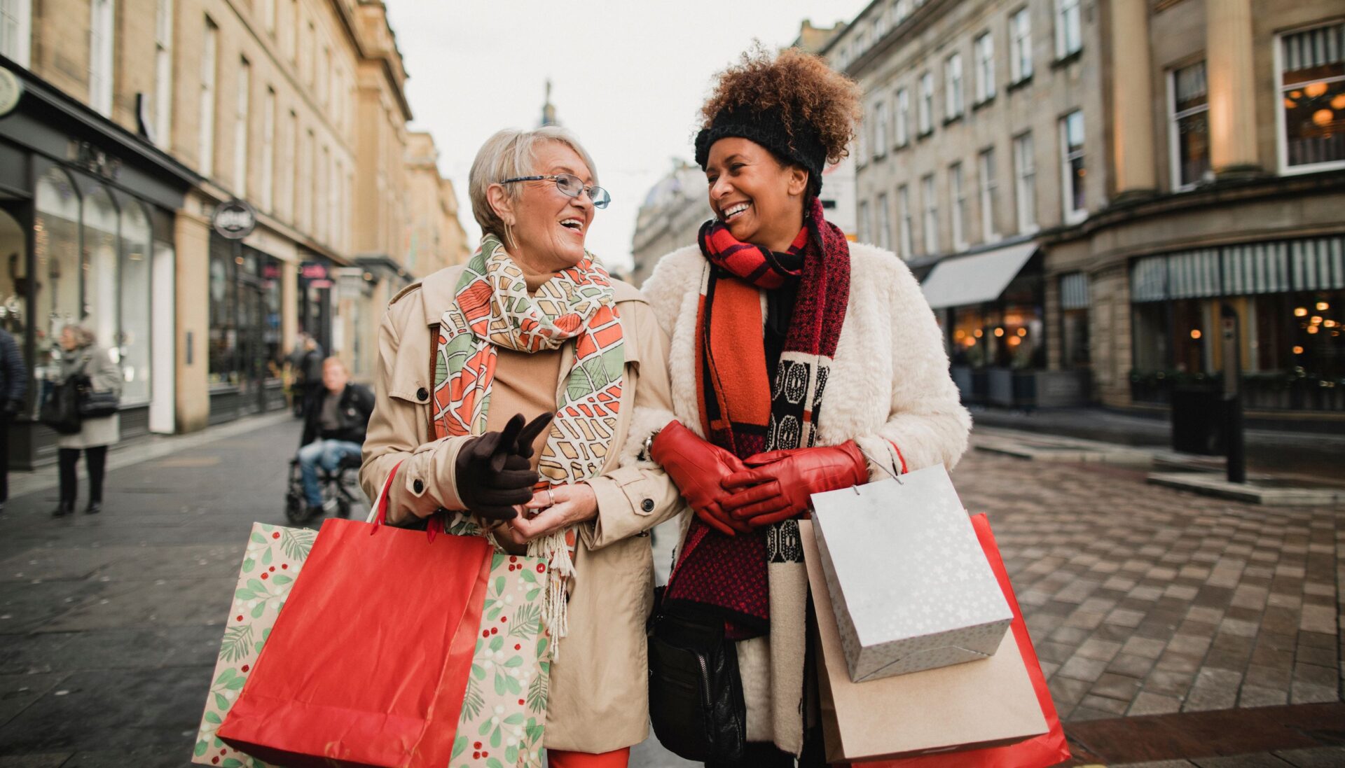 Two women with linked arms and dressed in reds and golds walking down a high street and laughing as they clutch Christmas bags of shopping